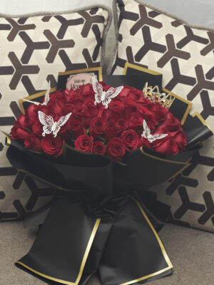 Ramo buchón de 100 rosas rojas con accesorios de mariposas y corona, envuelto en papel coreano negro con detalles dorados, arreglo floral lujoso visto de frente.