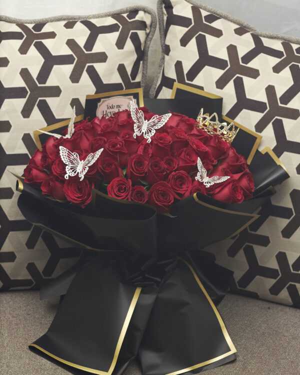 ramo_buchon_de_100_rosas_1 Ramo buchón de 100 rosas rojas con accesorios de mariposas y corona, envuelto en papel coreano negro con detalles dorados, arreglo floral lujoso visto de frente.
