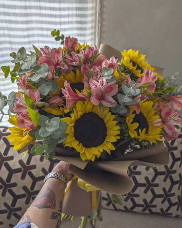Persona sosteniendo ramo con mix floral de girasoles, astromelias y mini dólar, arreglo natural y colorido listo para entregar.