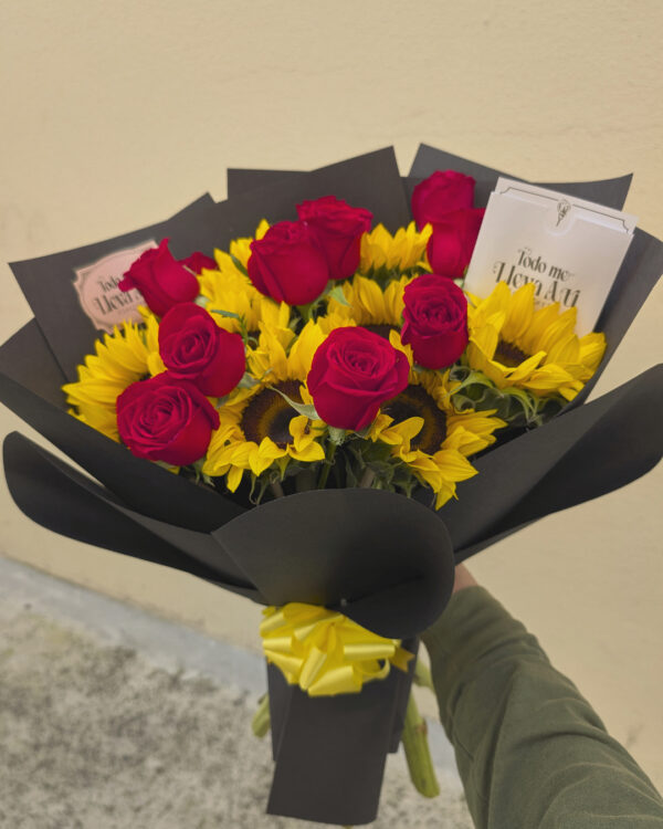 Persona sosteniendo ramo de girasoles y rosas rojas envuelto en papel negro, arreglo floral listo para entregar
