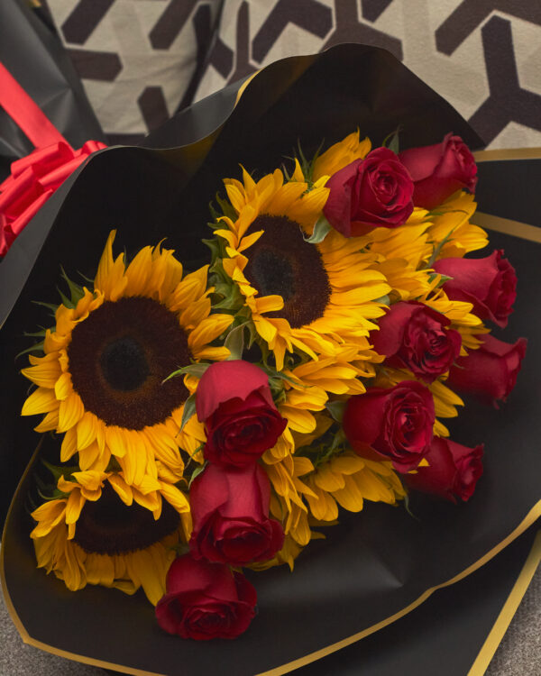 ramo_de_girasoles_y_rosas_rojas_con_papel_coreano_3 Detalle cercano de girasoles y rosas rojas en ramo con papel coreano negro, mostrando textura y contraste de colores.