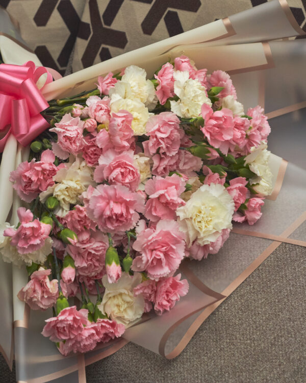 Detalle cercano de claveles blancos y rosas en ramo con envoltura de papel coreano y moño rosa, mostrando textura y suavidad floral.