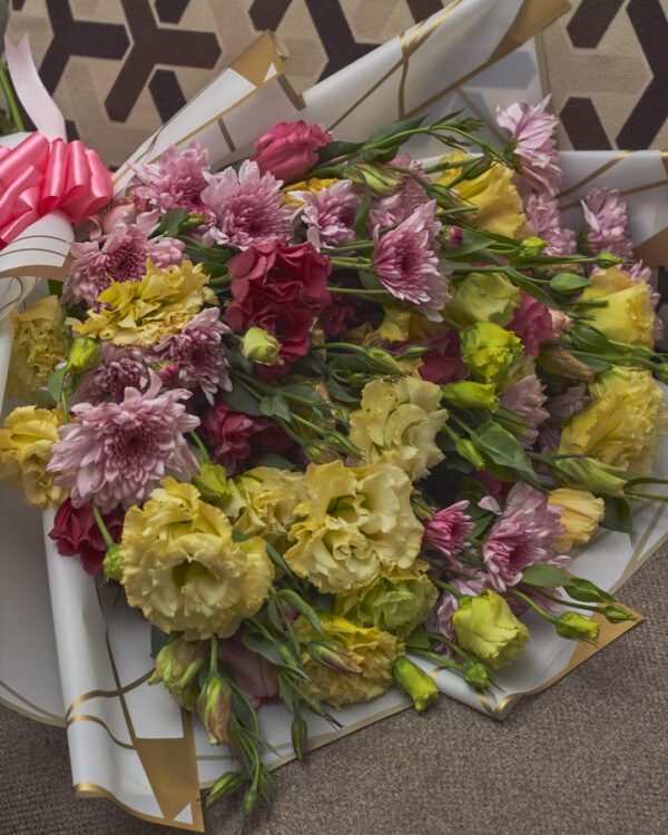 Detalle cercano de lisianthus amarillo y vino con crisantemos lilas malibú en ramo envuelto en papel coreano blanco y moño rosado, mostrando textura y contraste floral.