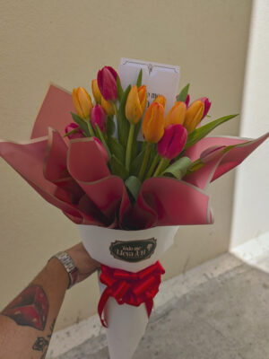 Persona sosteniendo ramo de tulipanes anaranjados y rojos, envuelto con papel coreano rojo y blanco, decorado con moño rojo, arreglo floral vibrante y elegante.