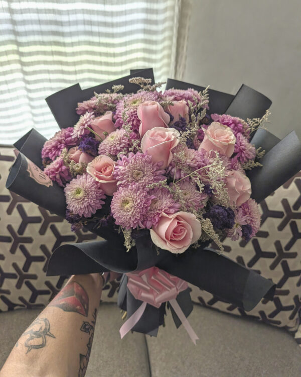 ramo_de_rosas_crisantemos_caspia_statice_3 Persona sosteniendo ramo redondo con rosas, crisantemos lilas, caspia y flor statice, envuelto en papel negro.