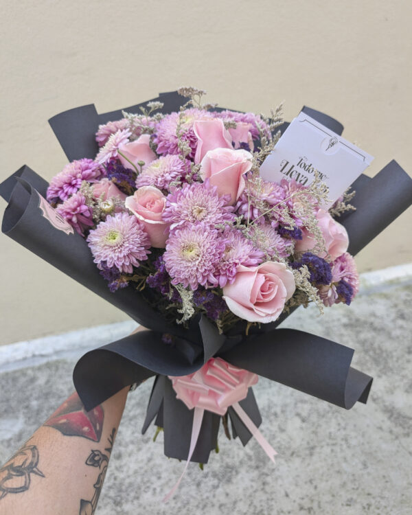 ramo_de_rosas_crisantemos_caspia_statice_5 Persona sosteniendo ramo redondo con rosas, crisantemos lilas, caspia y flor statice, envuelto en papel negro, fotografía tomada en exteriores.